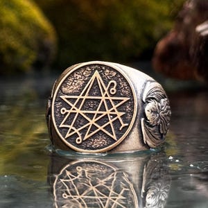May include: A silver-toned metal ring featuring a circular face with a textured, dark background and a raised, intricate geometric star symbol. The sides of the ring display detailed, sculpted relief carvings of wings, creating a bold, metallic aesthetic.