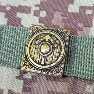 May include: A gold-coloured metal shield with a raised arrow design. The shield is attached to a green and brown camouflage fabric.