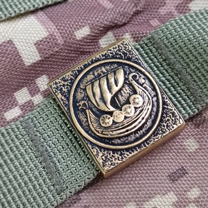 May include: A brass button with a detailed image of a Viking ship with oars and shields. The button is square and has a textured surface.