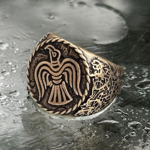 May include: A bronze-toned metal ring featuring a raised relief of a stylized bird with outstretched wings centered within a circular border. The sides of the ring band are decorated with intricate, swirling knotwork patterns in a dark finish.