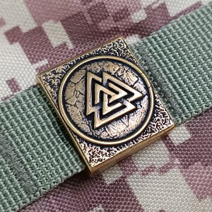 May include: A square metal charm with a textured surface. The charm features a circular design with a gold-coloured Celtic knot symbol inside. The charm is a popular symbol of protection and strength.