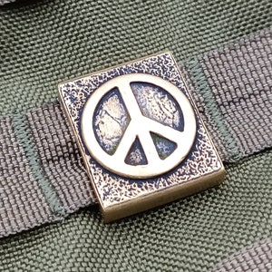 Op de afbeelding: Een messing vredestekenbedel met een getextureerd oppervlak. De bedel is bevestigd aan een groene en bruine stoffen achtergrond.