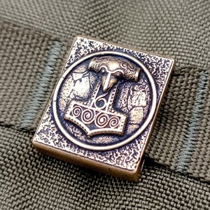 Może przedstawiać: Kwadratowy metalowy brelok w kolorze brązu z wypukłym okrągłym wzorem przedstawiającym stylizowany młotek z ptasim łbem na górze. Młotek jest otoczony ozdobną ramką.