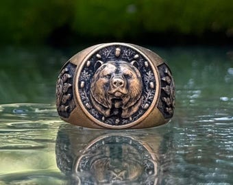 Könnte beinhalten: Ein bronzefarbener Metallring mit einer kreisförmigen Vorderseite, die ein detailliertes Relief eines Bärenkopfes zeigt, umgeben von einem Rand aus kleinen Punkten und blattartigen Mustern. Die Seiten des Rings weisen weitere strukturierte Elemente auf.
