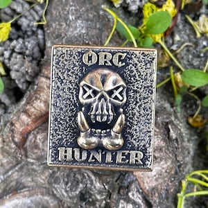 Könnte beinhalten: Ein quadratischer, bronzefarbener Metallpin mit den Worten "ORC HUNTER" und einem Totenkopf-Design. Der Schädel hat durchgestrichene Augen und große Stoßzähne. Der Pin ist vor einem strukturierten Hintergrund mit grünen Blättern und gelben Blüten platziert.