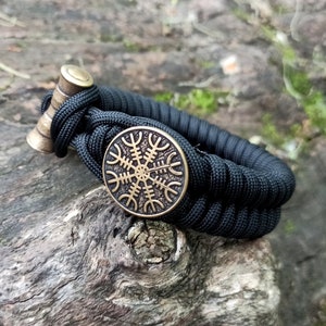 May include: Black paracord bracelet with a brass button and a round brass charm featuring a Vegvisir symbol.