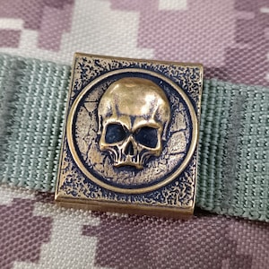 以下が含まれることがあります： 詳細なデザインが施された金色の金属製の頭蓋骨チャーム。頭蓋骨は、テクスチャ加工された背景を持つ円形のフレームにセットされています。チャームは緑色の布製のストラップに取り付けられています。