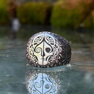 May include: A silver-toned metal ring featuring a detailed relief of a split face design, with one side showing a smooth profile and the other side displaying intricate knotwork patterns. The band has a textured, dark, oxidised finish throughout.