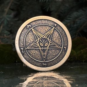 May include: A circular bronze-toned medallion featuring a central embossed pentagram with a goat head design. The outer ring contains five distinct Hebrew characters engraved at regular intervals. The surface has a textured, weathered finish.