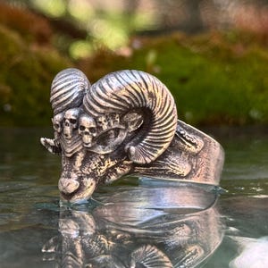 Pode incluir: Um anel de metal prateado com um design detalhado de cabeça de carneiro com grandes chifres curvos. A testa do carneiro é adornada com uma série de pequenas caveiras humanas esculpidas integradas no metal, criando um acabamento escuro.