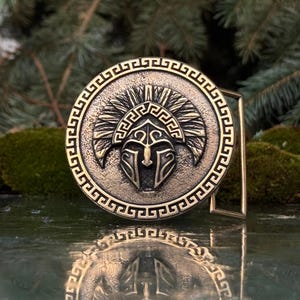 May include: A circular bronze-toned belt buckle featuring a central embossed Spartan helmet design. The outer border is decorated with a repeating Greek key pattern. The metal surface has a textured, weathered finish with dark recessed details.
