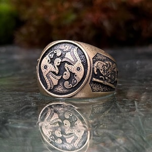 Op de afbeelding: Een bronskleurige metalen ring met een cirkelvormig voorvlak met twee in reliëf aangebrachte, gestileerde vogelachtige figuren die tegenover elkaar staan op een donkere, getextureerde achtergrond. De zijkant toont een extra gegraveerde figuur met een schild en speer.