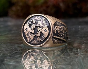May include: A bronze-toned metal ring featuring a circular face with two embossed, stylized bird-like figures facing each other against a dark, textured background. The side of the ring displays an additional engraved figure with a shield and spear.