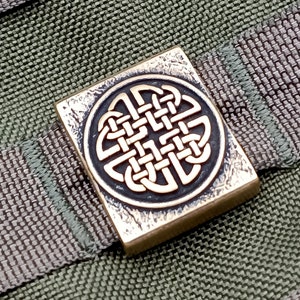 Peut inclure: Un charme en métal de couleur bronze de forme carrée avec un motif de nœud celtique. Le charme est sur un fond de tissu vert et gris.