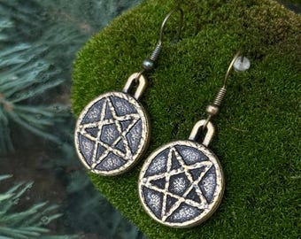 May include: A pair of dangle earrings featuring circular bronze-toned pendants with a raised five-pointed star design. The metal has a dark, textured finish within the recessed areas, and each earring hangs from a simple curved metal hook wire.
