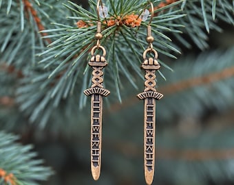 May include: A pair of bronze-toned metal earrings shaped like long swords. Each earring features a detailed hilt with a cross-guard and a blade engraved with the text LIEFBERHT in a vertical orientation, suspended from a simple hook wire.