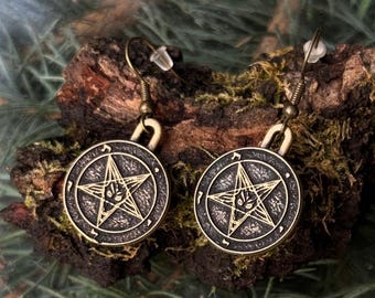 May include: A pair of bronze-toned metal drop earrings featuring a circular pendant with a raised pentagram design. Each earring has a central star motif with an animal-like face at the center, suspended from a simple curved metal hook wire.