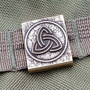 Peut inclure: Un charme carré en métal argenté avec un motif de nœud celtique. Le nœud est au centre du charme et est entouré d'un cercle. Le charme a une surface texturée.