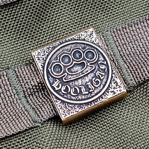 Könnte beinhalten: Ein quadratischer Schlüsselanhänger aus Messing mit einem erhabenen Bild eines Schlagringes und dem Wort "HOOLIGAN" in gotischer Schrift.