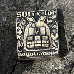 May include: A gold-colored metal pin with a camouflage pattern and the text "SUIT for negotiations". The pin features a stylized image of a body armor vest with the Ukrainian coat of arms.