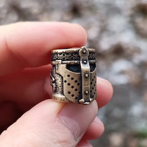 May include: A small, detailed, bronze-colored metal charm in the shape of a knight's helmet. The helmet has a decorative pattern around the top and a faceplate with a mesh design.