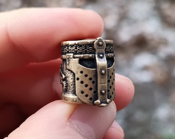 May include: A small, detailed, bronze-colored metal charm in the shape of a knight's helmet. The helmet has a decorative pattern around the top and a faceplate with a mesh design.