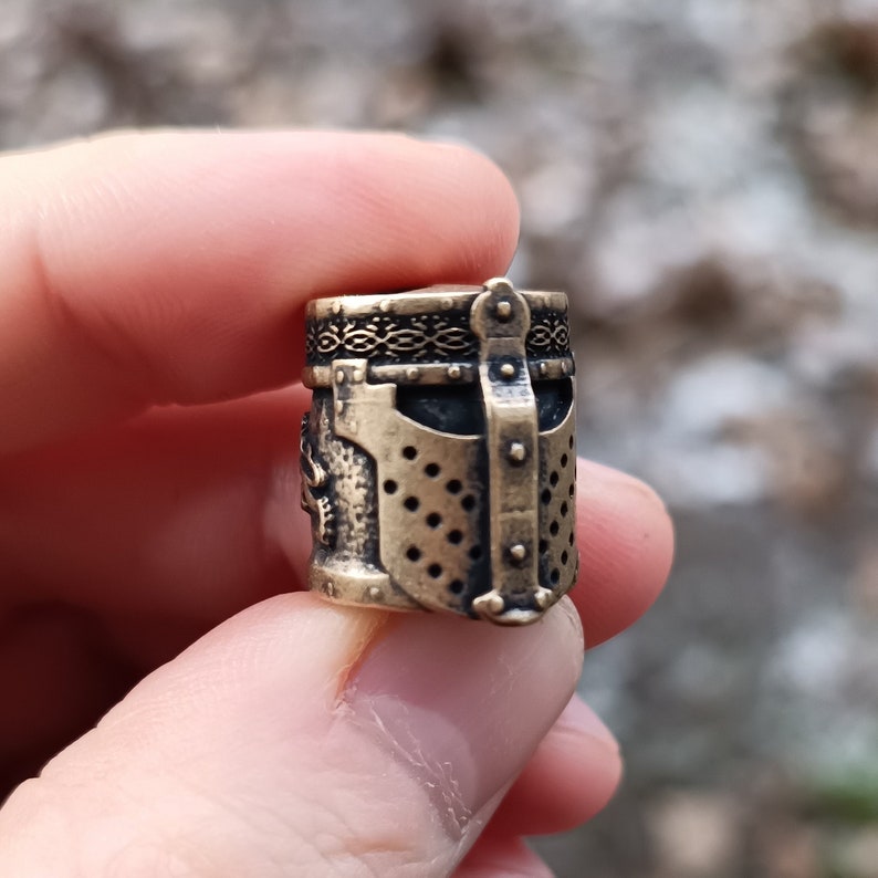 May include: A small, detailed, bronze-colored metal charm in the shape of a knight's helmet. The helmet has a decorative pattern around the top and a faceplate with a mesh design.