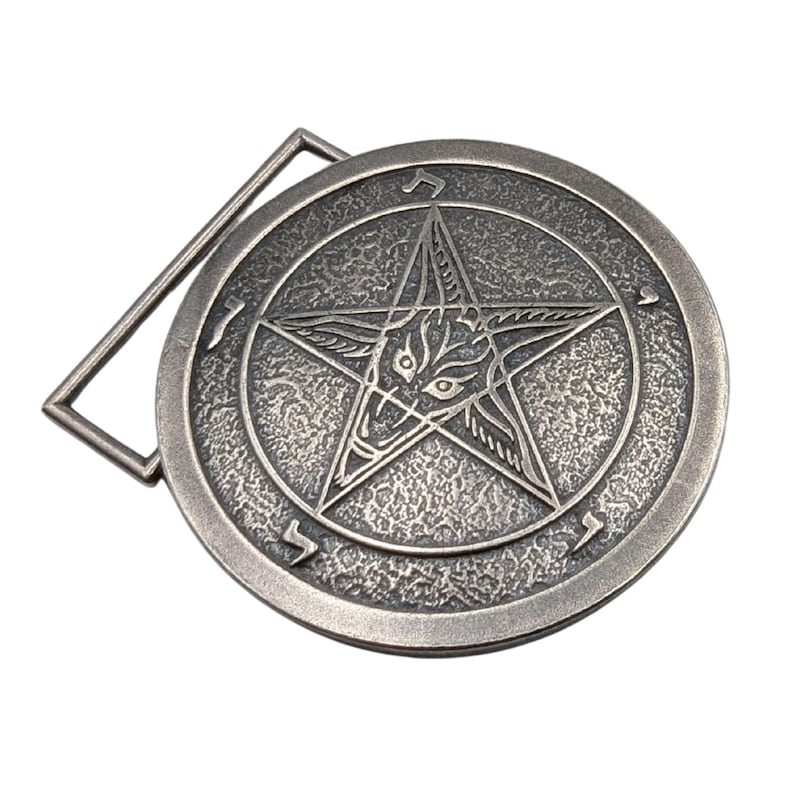 May include: A circular silver-toned metal belt buckle featuring a raised pentagram design with a central goat head motif. The background of the circle has a textured, hammered finish, and Hebrew characters are engraved along the outer edge.