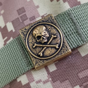 Könnte beinhalten: Ein quadratischer, goldfarbener Metallstift mit einem Totenkopf- und Knochenkreuz-Motiv. Der Stift ist an einem grün-braunen Tarnstoff befestigt.
