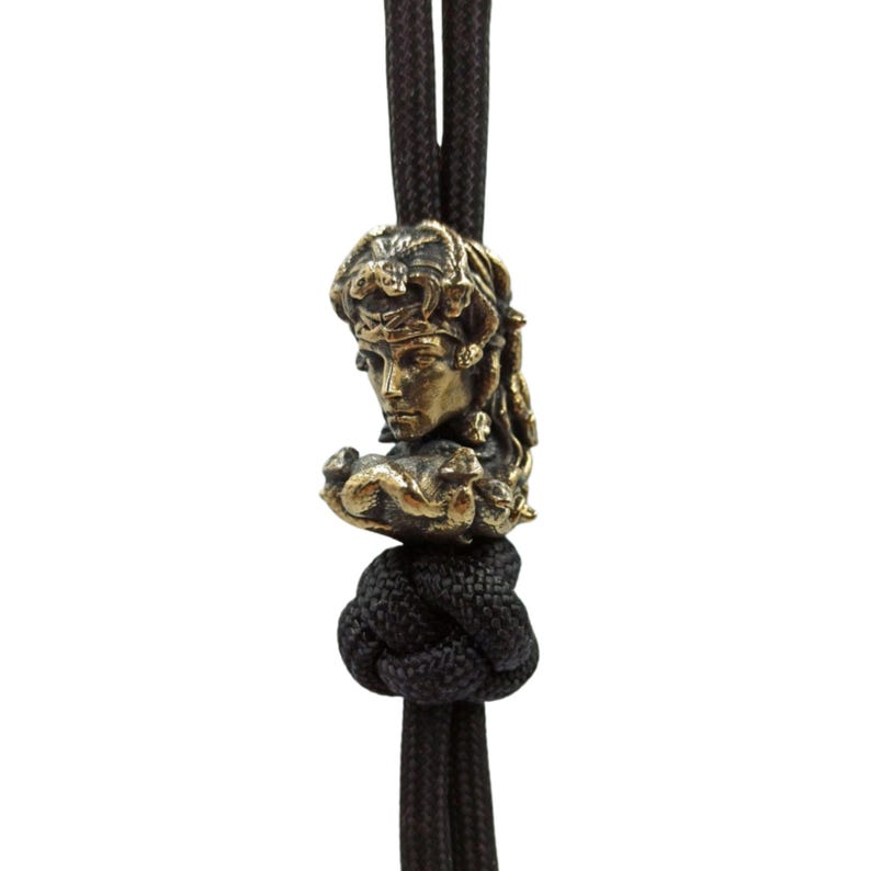Puede incluir: Una cuenta de metal de color dorado con una escultura detallada de la cara de una mujer con cabello y follaje intrincados. La cuenta est&aacute; unida a un cord&oacute;n negro.
