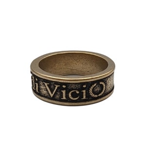 May include: A bronze-toned metal ring with a textured, darkened band featuring the embossed text "Veni Vidi Vici" and a small laurel wreath symbol. The ring has a smooth, polished top edge and a wide, flat profile with a weathered, antique finish.