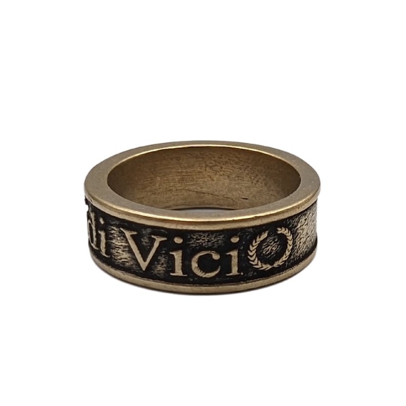 May include: A bronze-toned metal ring with a textured, darkened band featuring the embossed text "Veni Vidi Vici" and a small laurel wreath symbol. The ring has a smooth, polished top edge and a wide, flat profile with a weathered, antique finish.