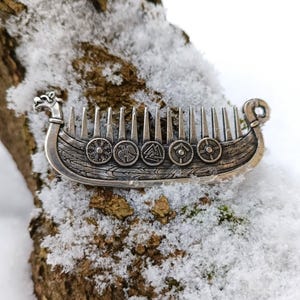 A silver-colored Viking ship Drakkar comb with intricate details. The comb features a ship design with shields and a dragon head at the front. The comb is resting on a snow-covered woody surface.