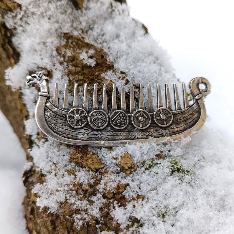 A silver-colored Viking ship Drakkar comb with intricate details. The comb features a ship design with shields and a dragon head at the front. The comb is resting on a snow-covered woody surface.