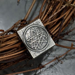 May include: A rectangular silver-toned metal pendant with a weathered, textured surface. The center features a circular emblem containing an intricate seven-pointed star design with small circular terminals at the points of the star lines.