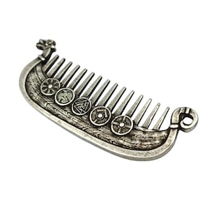 A silver plated beard comb shaped like a Viking longship Drakkar. The comb features detailed carvings of shields and Viking symbols along the ship's hull and on the comb's handle. The comb has a decorative norse design.