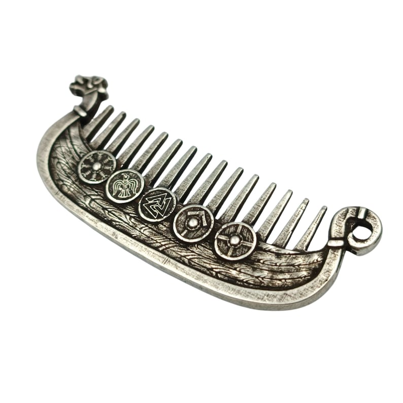 A silver plated beard comb shaped like a Viking longship Drakkar. The comb features detailed carvings of shields and Viking symbols along the ship's hull and on the comb's handle. The comb has a decorative norse design.
