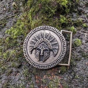 May include: A circular metal belt buckle with a bronze-toned finish featuring a raised relief of a crested helmet in the centre. The design is surrounded by a decorative Greek key border pattern and includes a rectangular loop on the right side.