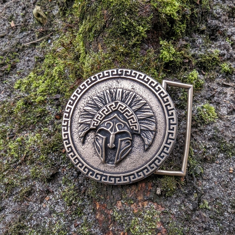 May include: A circular metal belt buckle with a bronze-toned finish featuring a raised relief of a crested helmet in the centre. The design is surrounded by a decorative Greek key border pattern and includes a rectangular loop on the right side.