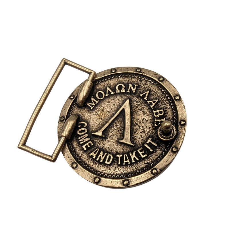 May include: A circular brass-toned belt buckle featuring a raised Greek Lambda symbol in the centre. The text "&Mu;&Omicron;&Lambda;&Omega;&Nu; &Lambda;&Alpha;&Beta;&Epsilon;" is inscribed above the symbol, and the English text "COME AND TAKE IT" is embossed along the bottom edge of the buckle.