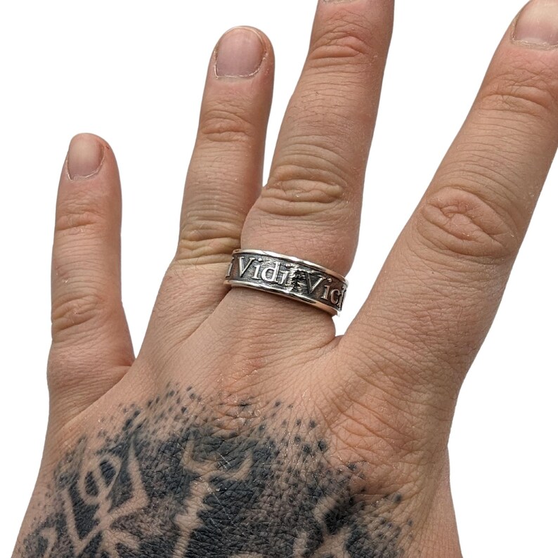 May include: A silver-toned metal band ring featuring a textured, oxidised finish. The surface of the ring is engraved with the Latin phrase Vidi Vici in a serif font, with the letters appearing dark against the polished silver metal background.