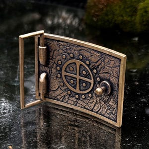 May include: A rectangular bronze-toned belt buckle featuring a textured, weathered surface. The center displays a circular emblem with a cross and dot pattern, surrounded by a ring of small raised dots. It includes a metal frame and a central stud.