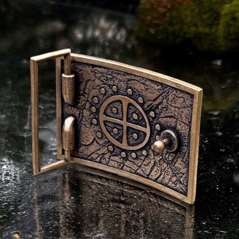 May include: A rectangular bronze-toned belt buckle featuring a textured, weathered surface. The center displays a circular emblem with a cross and dot pattern, surrounded by a ring of small raised dots. It includes a metal frame and a central stud.