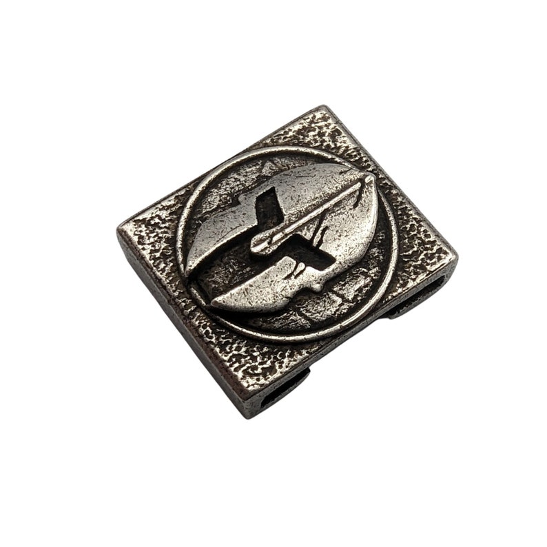 May include: A silver-toned metal charm with a textured surface. The charm features a circular design with a stylized image of a shield and a sword.
