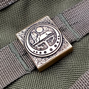 May include: A brass-coloured metal buckle with a detailed design of a stylised animal lying down inside a circle. The buckle has a textured surface.