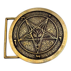 May include: A circular brass belt buckle featuring a raised pentagram design with a central goat head motif. The background of the circular plate has a textured, weathered finish. Hebrew characters are engraved along the outer edge of the circle.