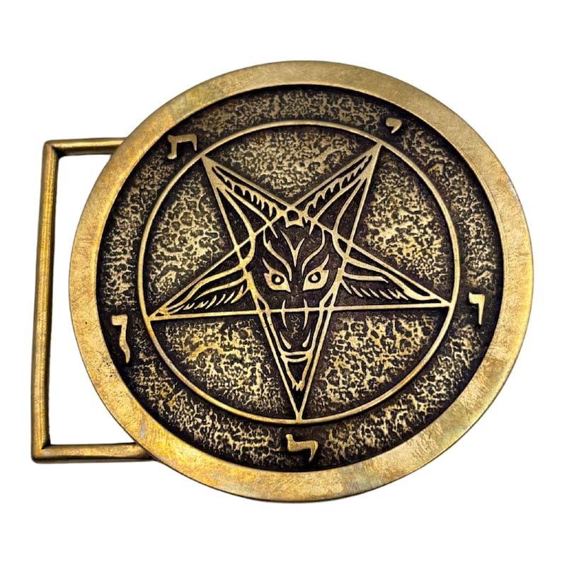 May include: A circular brass belt buckle featuring a raised pentagram design with a central goat head motif. The background of the circular plate has a textured, weathered finish. Hebrew characters are engraved along the outer edge of the circle.