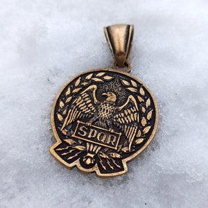 May include: A circular bronze-toned pendant featuring a relief of an eagle with wings spread, clutching a banner inscribed with the letters SPQR. The eagle is framed by a laurel wreath, with lightning bolts depicted at the base of the design.