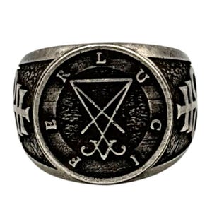 May include: A silver-toned metal ring featuring a circular face with a textured dark background. The center displays a sigil of Lucifer, surrounded by the letters L, U, C, I, F, E, R. The sides of the ring are adorned with embossed cross symbols.