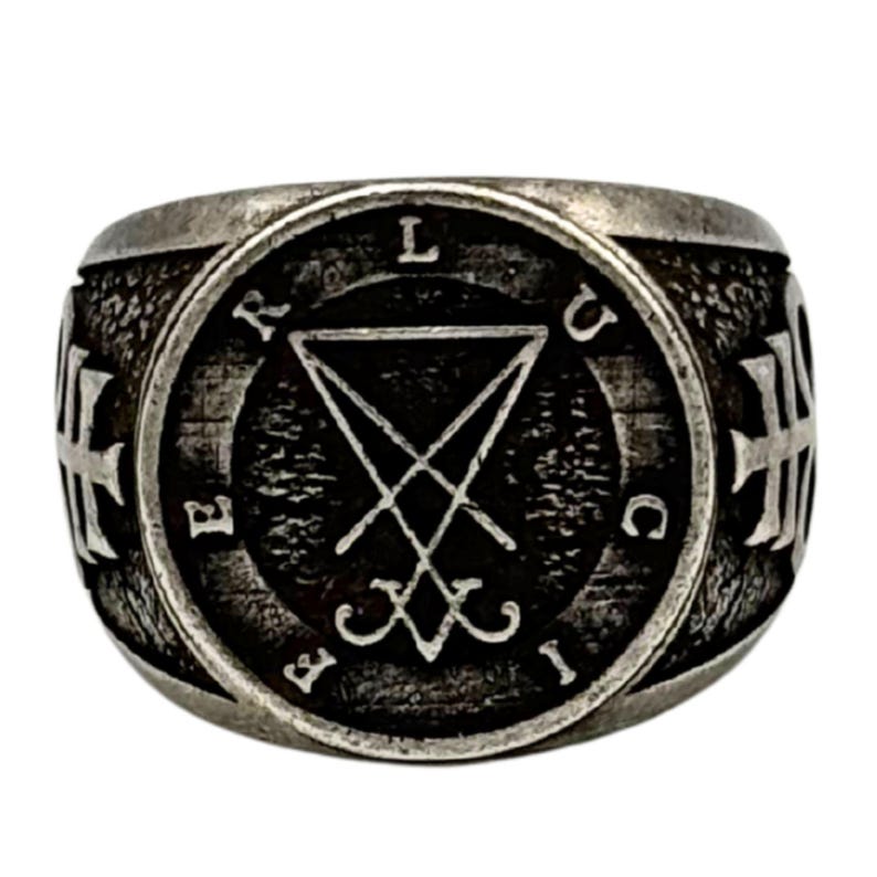 May include: A silver-toned metal ring featuring a circular face with a textured dark background. The center displays a sigil of Lucifer, surrounded by the letters L, U, C, I, F, E, R. The sides of the ring are adorned with embossed cross symbols.